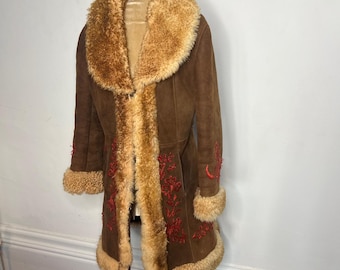 Puede incluir: Una larga chaqueta de ante marrón con cuello y forro de piel sintética. La chaqueta tiene bordados rojos en la parte delantera y las mangas.
