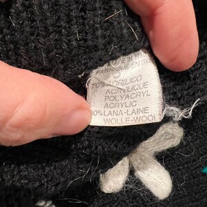 May include: Black knitted fabric with white and teal embroidered flowers. The fabric label reads "Made in Italy, Fab. in Italia, 70% Acrilico Acrylic Polyacryl, 30% Lana-Laine Wolle-Wool".
