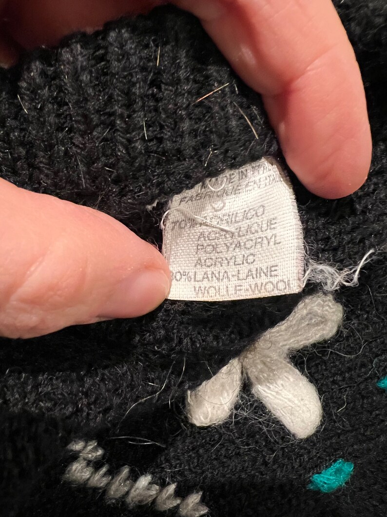 May include: Black knitted fabric with white and teal embroidered flowers. The fabric label reads "Made in Italy, Fab. in Italia, 70% Acrilico Acrylic Polyacryl, 30% Lana-Laine Wolle-Wool".