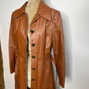 May include: A vintage, long, brown leather coat with a button-up front and a belt. The coat is displayed on a mannequin and has a classic, tailored design. The coat's color is a rich, warm brown.