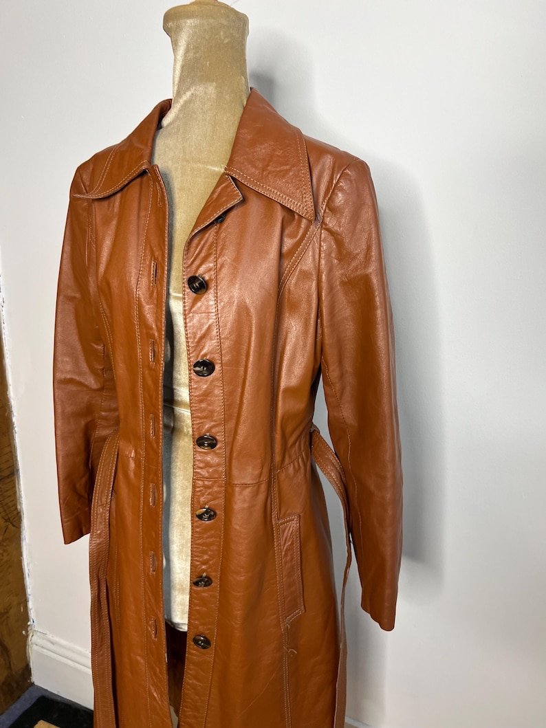 May include: A vintage, long, brown leather coat with a button-up front and a belt. The coat is displayed on a mannequin and has a classic, tailored design. The coat's color is a rich, warm brown.
