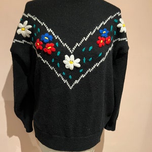 May include: Black knitted sweater with a white, green, and blue embroidered floral design on the front.