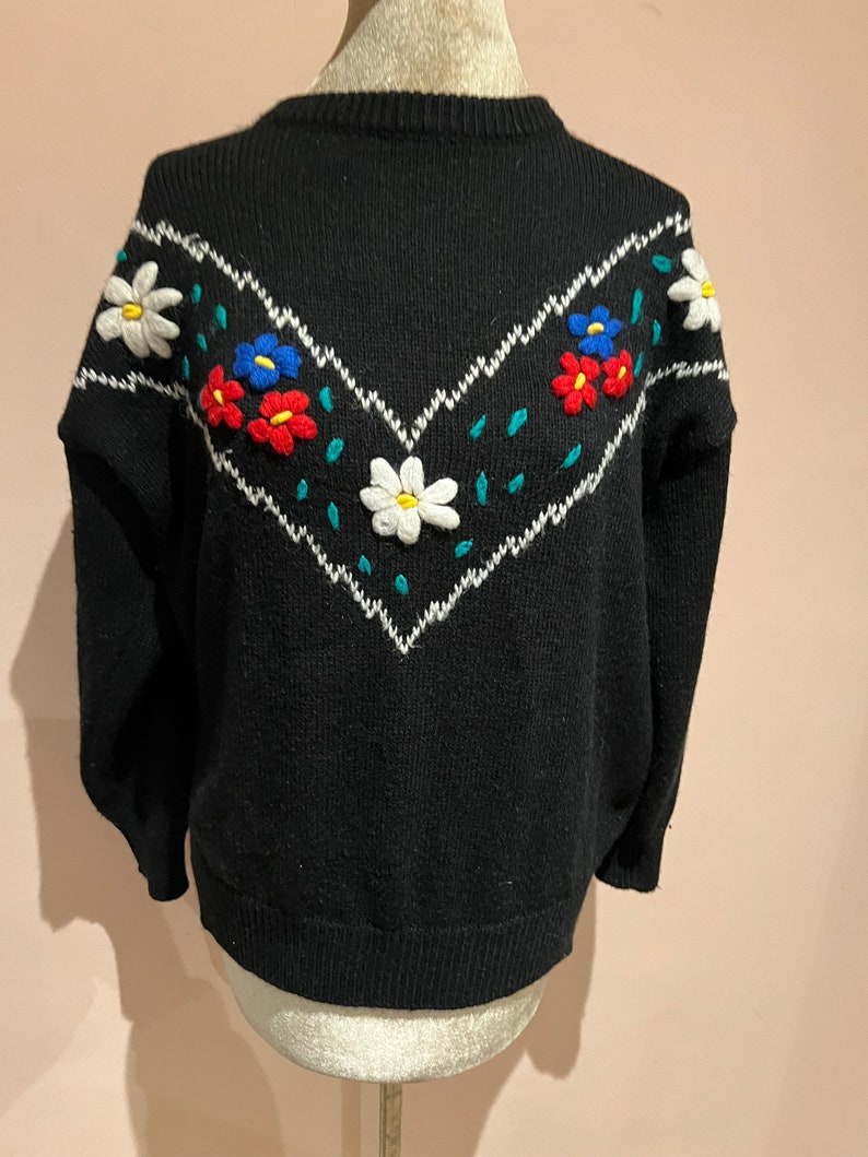 May include: Black knitted sweater with a white, green, and blue embroidered floral design on the front.
