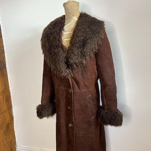 May include: A long, brown suede coat with a dark brown fur collar and cuffs. The coat is buttoned down the front and appears to be vintage. The coat is displayed on a mannequin.