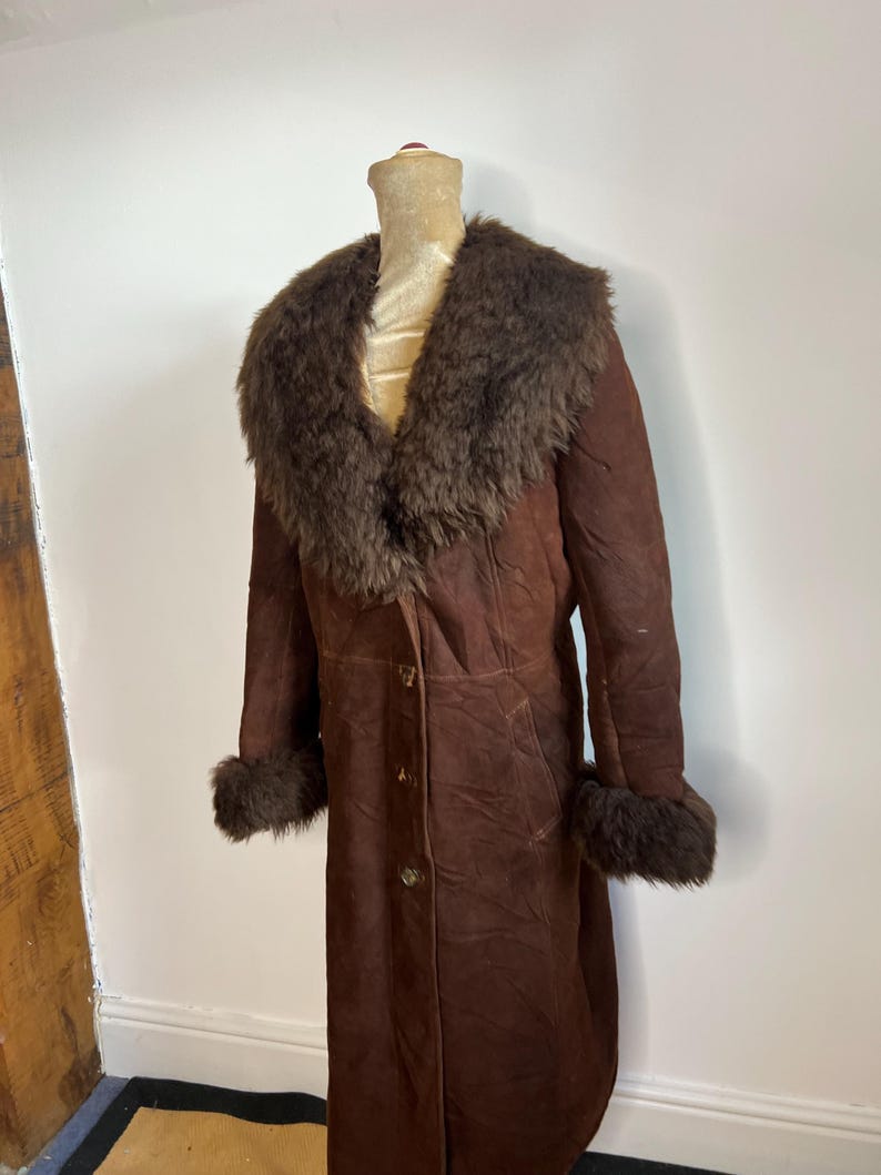 May include: A long, brown suede coat with a dark brown fur collar and cuffs. The coat is buttoned down the front and appears to be vintage. The coat is displayed on a mannequin.