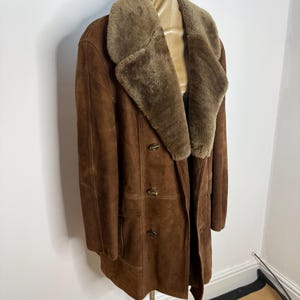 May include: A brown suede coat with a shearling collar and lining. The double-breasted coat has two rows of buttons and two front pockets. The coat is displayed on a mannequin. The coat's length is approximately 102 cm.