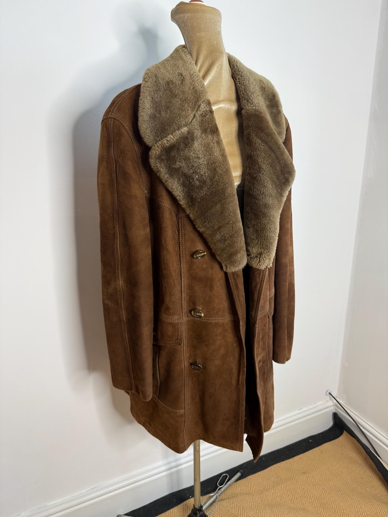 May include: A brown suede coat with a shearling collar and lining. The double-breasted coat has two rows of buttons and two front pockets. The coat is displayed on a mannequin. The coat's length is approximately 102 cm.