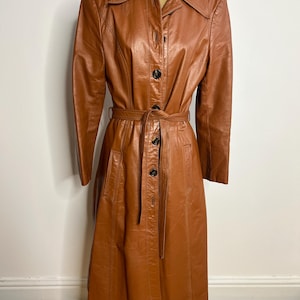 May include: A vintage brown leather trench coat with a button-up front, a belt, and a collared neckline. The coat is displayed on a mannequin and has a classic, tailored design. The coat is a warm, medium brown color.