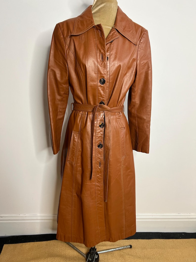 May include: A vintage brown leather trench coat with a button-up front, a belt, and a collared neckline. The coat is displayed on a mannequin and has a classic, tailored design. The coat is a warm, medium brown color.