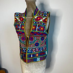 May include: A colorful, embroidered vest with a teal background and intricate floral and geometric patterns in red, yellow, blue, and white. The vest is sleeveless with a high collar and is displayed on a mannequin.