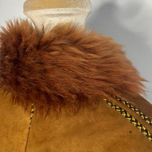 May include: Close-up of a brown suede jacket with a faux fur collar. The jacket features decorative stitching in black and yellow along the seams. The collar is a rich, reddish-brown color, adding a touch of warmth to the garment.