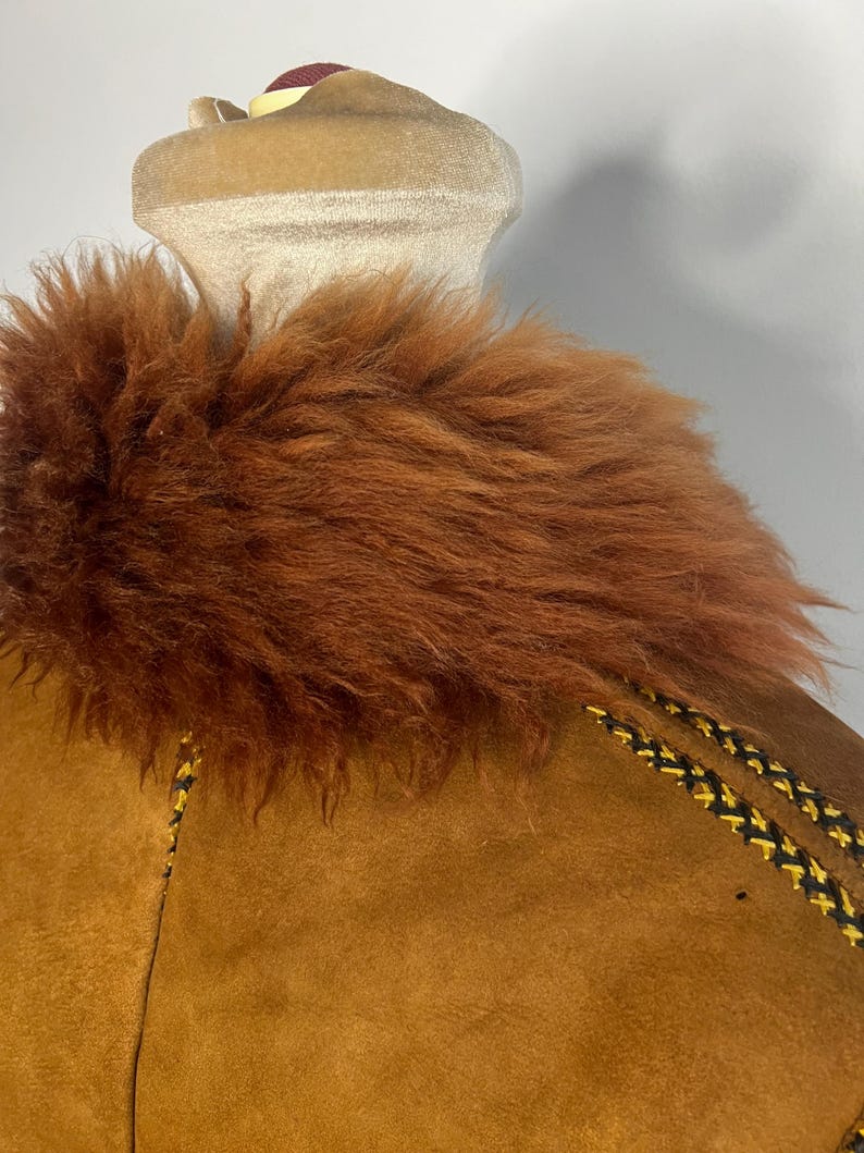 May include: Close-up of a brown suede jacket with a faux fur collar. The jacket features decorative stitching in black and yellow along the seams. The collar is a rich, reddish-brown color, adding a touch of warmth to the garment.