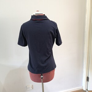 May include: A navy blue short-sleeved polo shirt on a mannequin. The shirt has a red and white collar.