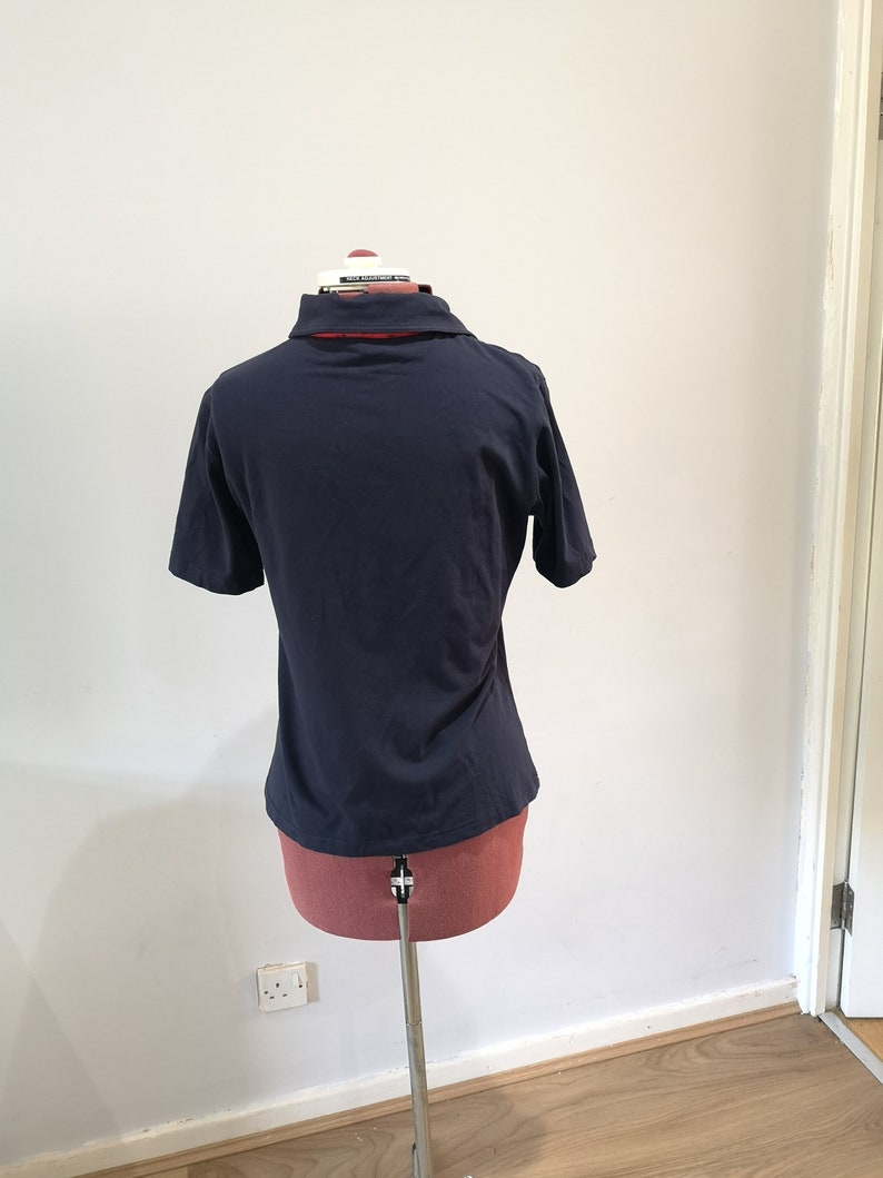 May include: A navy blue short-sleeved polo shirt on a mannequin. The shirt has a red and white collar.