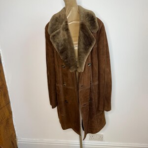 May include: A brown suede coat with a shearling collar and lining. The double-breasted coat features button closures and pockets. The coat is displayed on a mannequin, highlighting its full length and classic design.