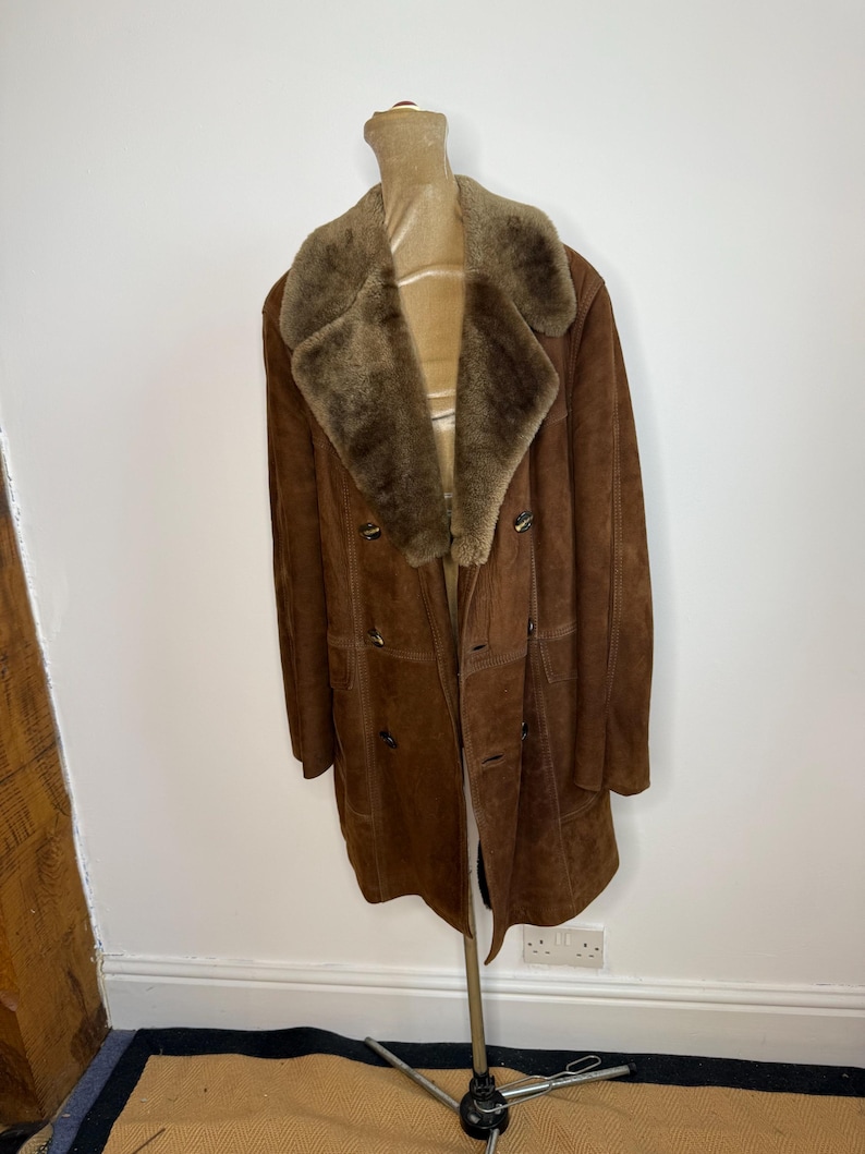 May include: A brown suede coat with a shearling collar and lining. The double-breasted coat features button closures and pockets. The coat is displayed on a mannequin, highlighting its full length and classic design.