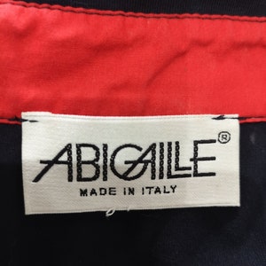 May include: White label with black text that reads "ABIGAILE" with a registered trademark symbol and "MADE IN ITALY" below the brand name. The label is attached to a black fabric.