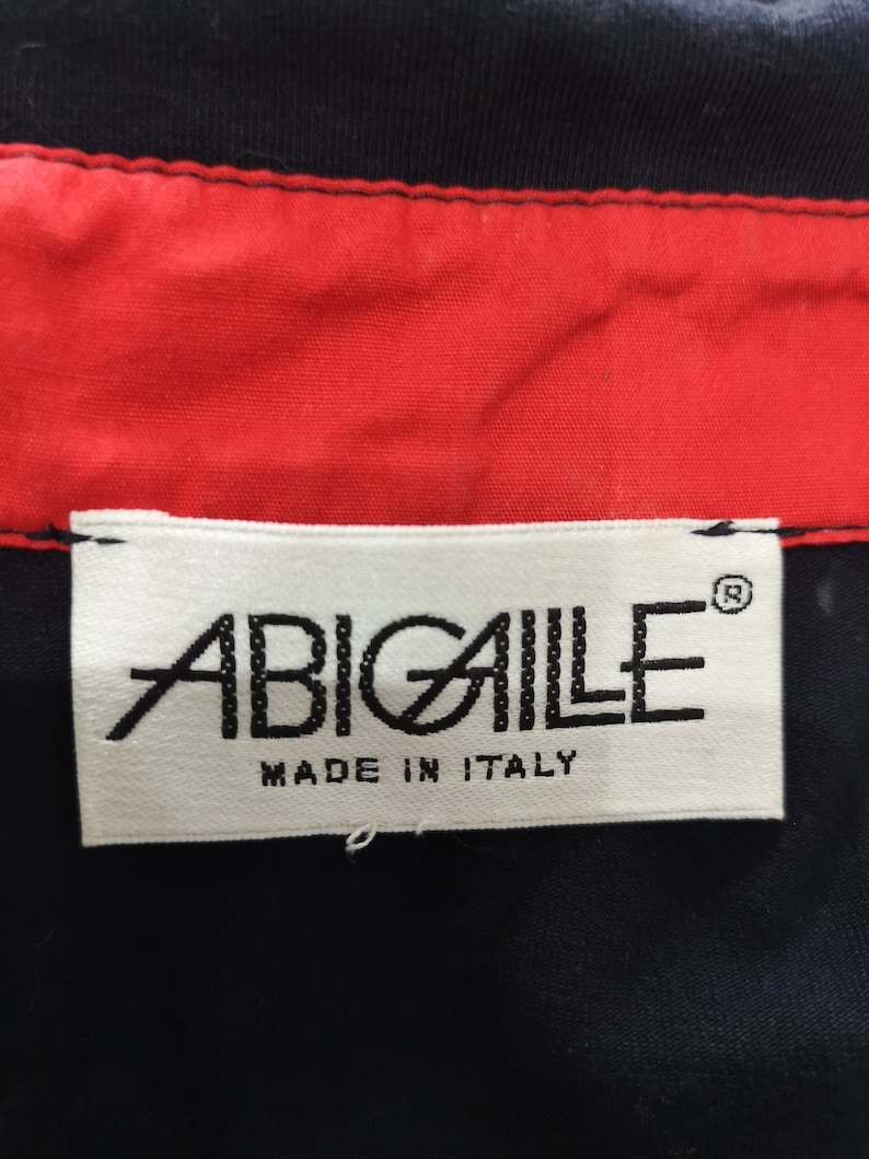 May include: White label with black text that reads "ABIGAILE" with a registered trademark symbol and "MADE IN ITALY" below the brand name. The label is attached to a black fabric.