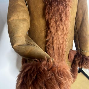 May include: A brown suede coat with a reddish-brown fur trim along the front and cuffs. The coat features a classic design with a warm, inviting aesthetic. The sleeves are long, and the coat appears to be well-constructed.