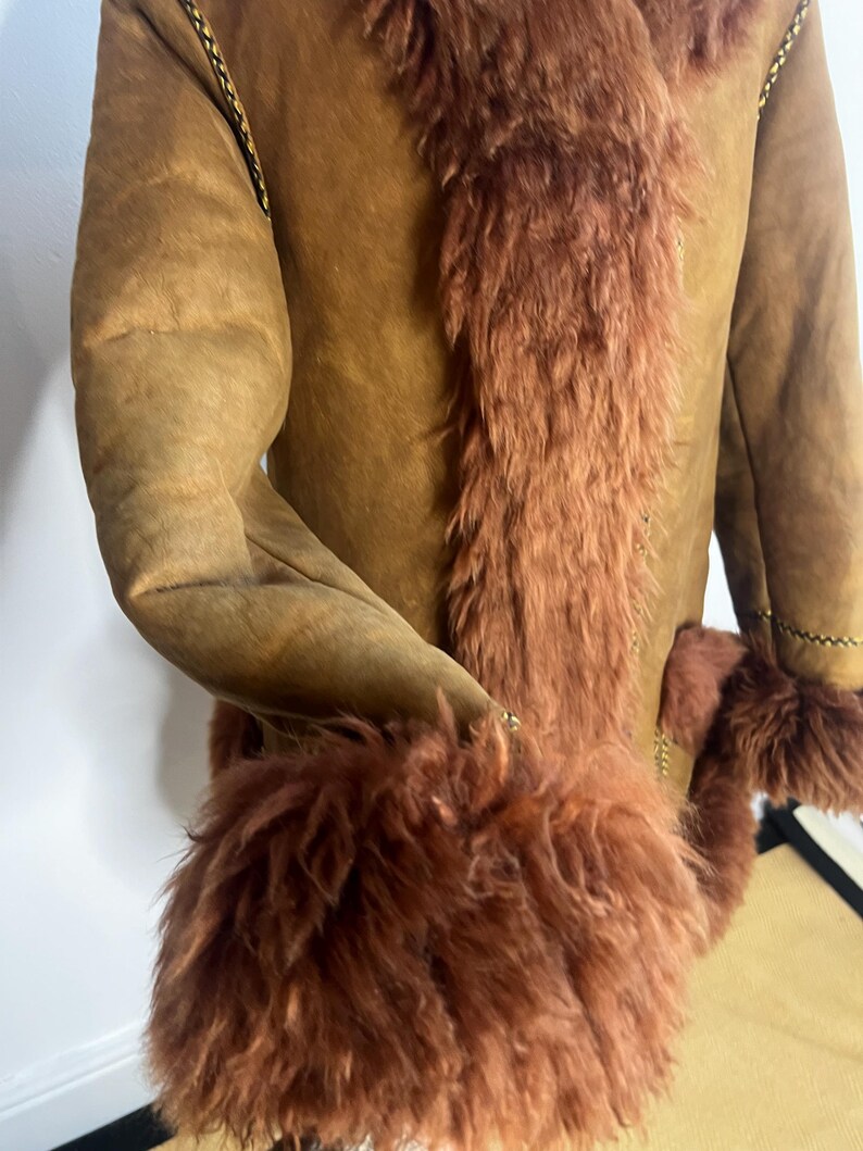 May include: A brown suede coat with a reddish-brown fur trim along the front and cuffs. The coat features a classic design with a warm, inviting aesthetic. The sleeves are long, and the coat appears to be well-constructed.