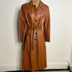 May include: A vintage brown leather coat displayed on a mannequin. The coat features a button-down front, a belt at the waist, and a collared neckline. The coat is knee-length and has long sleeves. The mannequin is on a metal stand.