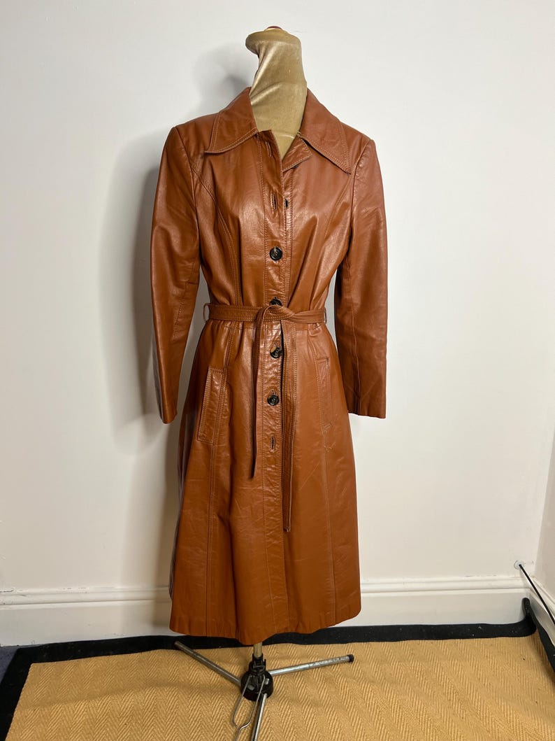 May include: A vintage brown leather coat displayed on a mannequin. The coat features a button-down front, a belt at the waist, and a collared neckline. The coat is knee-length and has long sleeves. The mannequin is on a metal stand.
