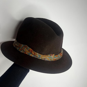 Könnte beinhalten: Dunkelbrauner Fedora-Hut mit einem gemusterten Band in Blau-, Orange- und Gelbtönen. Der Hut hat eine klassische Tränenkronenform und eine breite Krempe. Ein stilvolles Accessoire.
