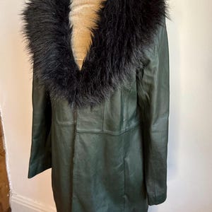 May include: A long, dark green leather coat with a black faux fur collar. The coat is displayed on a mannequin and has a classic, tailored design. The coat appears to be knee-length and has a button closure.