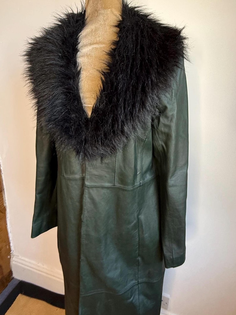 May include: A long, dark green leather coat with a black faux fur collar. The coat is displayed on a mannequin and has a classic, tailored design. The coat appears to be knee-length and has a button closure.