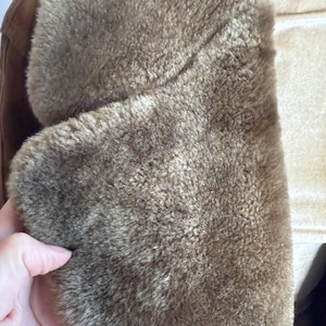 May include: Close-up of a brown shearling coat collar. The coat has a soft, textured appearance with a light brown colour. The collar is folded over, revealing the plush interior. The coat appears warm and cosy.