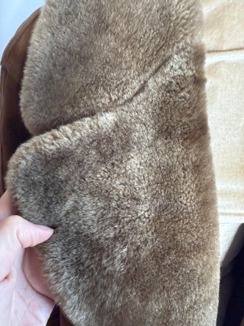 May include: Close-up of a brown shearling coat collar. The coat has a soft, textured appearance with a light brown colour. The collar is folded over, revealing the plush interior. The coat appears warm and cosy.