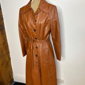 May include: A vintage brown leather trench coat with a belted waist and button-up front. The coat has a classic collar and long sleeves. The coat is displayed on a mannequin.