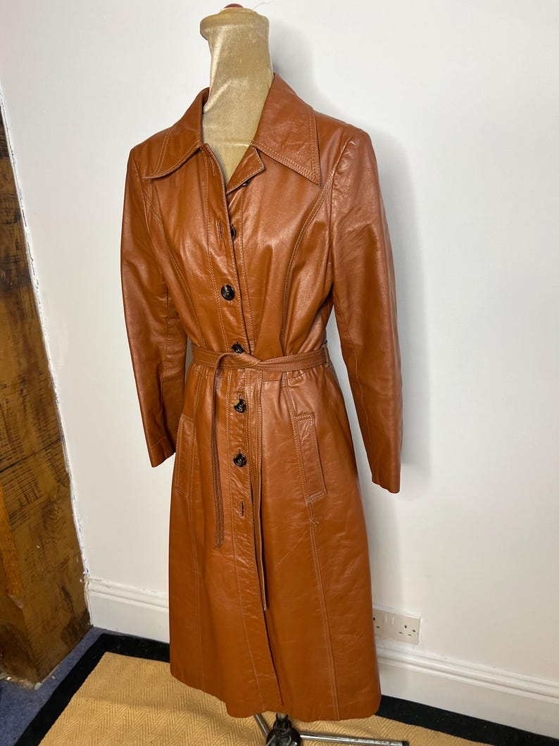 May include: A vintage brown leather trench coat with a belted waist and button-up front. The coat has a classic collar and long sleeves. The coat is displayed on a mannequin.