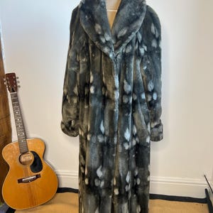May include: A long, faux fur coat in shades of gray and white, displayed on a mannequin. The coat has a wide collar and a pattern of irregular white spots. The coat is floor-length and appears to be vintage.