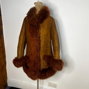 May include: A vintage brown suede coat with a faux fur collar, cuffs, and trim. The coat features a button-up front and decorative stitching. The coat is displayed on a mannequin.