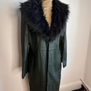 May include: A long, dark green coat with a black faux fur collar. The coat is displayed on a mannequin and appears to be made of leather or a leather-like material. The coat has a classic, elegant design.