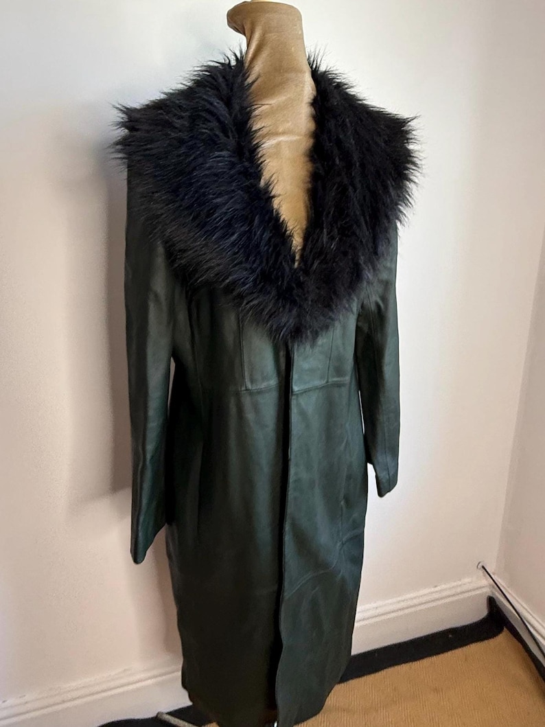 May include: A long, dark green coat with a black faux fur collar. The coat is displayed on a mannequin and appears to be made of leather or a leather-like material. The coat has a classic, elegant design.