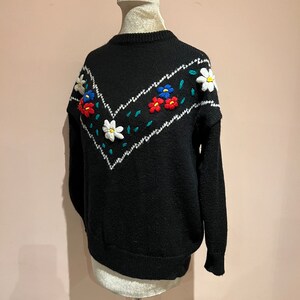 May include: Black knitted sweater with a white and green chevron pattern and embroidered flowers in red, blue, and white.