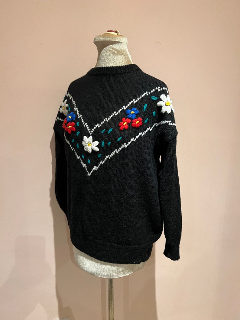 May include: Black knitted sweater with a white and green chevron pattern and embroidered flowers in red, blue, and white.