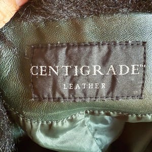 May include: Close-up of a dark green leather jacket with black faux fur trim. The label reads "CENTIGRADE" and "LEATHER" in white text on a black background. The jacket appears to be well-made and has a smooth texture.