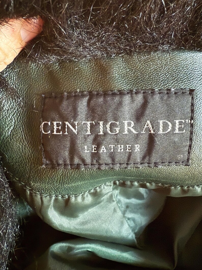 May include: Close-up of a dark green leather jacket with black faux fur trim. The label reads "CENTIGRADE" and "LEATHER" in white text on a black background. The jacket appears to be well-made and has a smooth texture.
