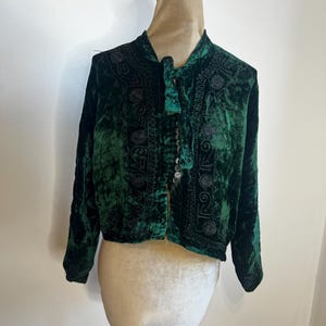 Może przedstawiać: Ciemnozielona aksamitna kurtka z misternymi haftami. Kurtka posiada wysoki kołnierz, długie rękawy i wiązanie. Tkanina ma pogniecioną fakturę, a kurtka jest prezentowana na manekinie.