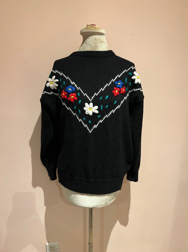 May include: Black knitted sweater with a white, red, blue, and green embroidered floral design.