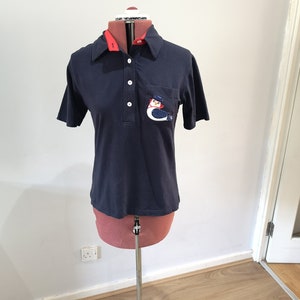 May include: A navy blue short-sleeved polo shirt with a red collar and a white button placket. The shirt has a pocket on the left chest with an embroidered bird design.