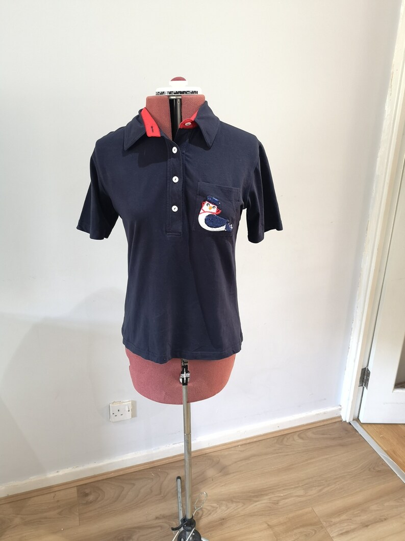 May include: A navy blue short-sleeved polo shirt with a red collar and a white button placket. The shirt has a pocket on the left chest with an embroidered bird design.