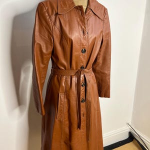 May include: A vintage brown leather trench coat with a button-up front, long sleeves, and a matching belt. The coat features a classic collar and two front pockets. The coat measures approximately 48 inches in length.