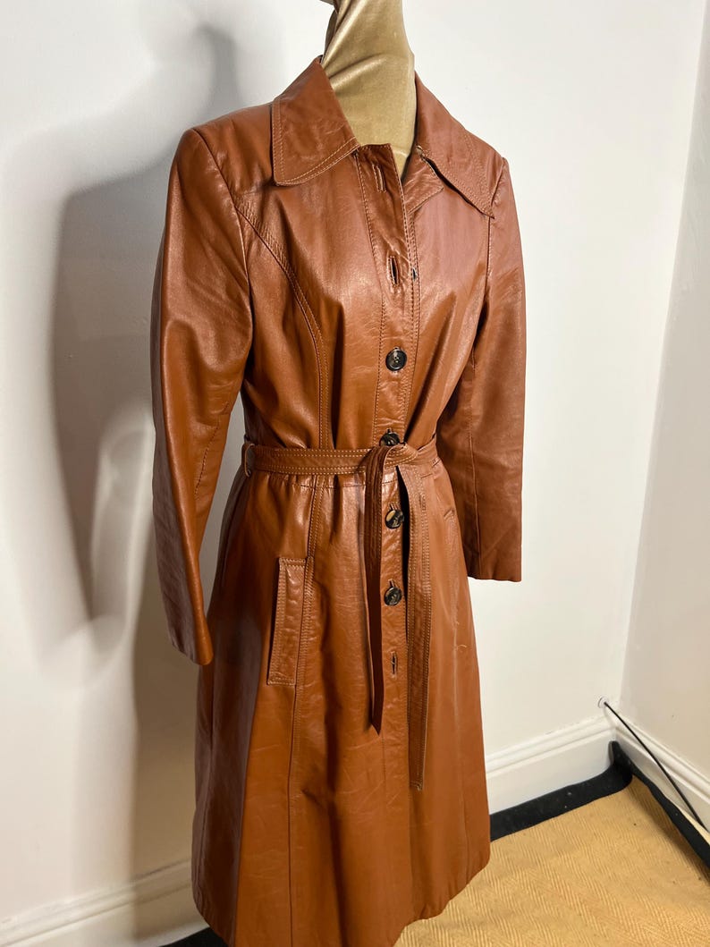 May include: A vintage brown leather trench coat with a button-up front, long sleeves, and a matching belt. The coat features a classic collar and two front pockets. The coat measures approximately 48 inches in length.
