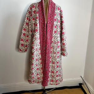 May include: A long, quilted robe with a floral design. The robe has a cream background with a repeating pattern of red flowers and green leaves. The trim and inner lining are a vibrant pink. The robe is displayed on a mannequin.