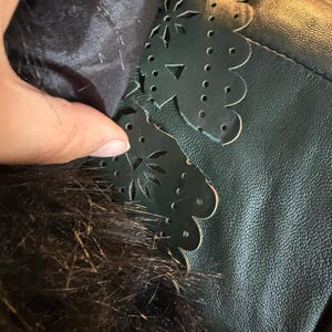 May include: Close-up of a dark green leather jacket detail. The jacket features a scalloped edge with floral cutouts. The interior lining is a dark fabric, and the collar has dark fur trim.