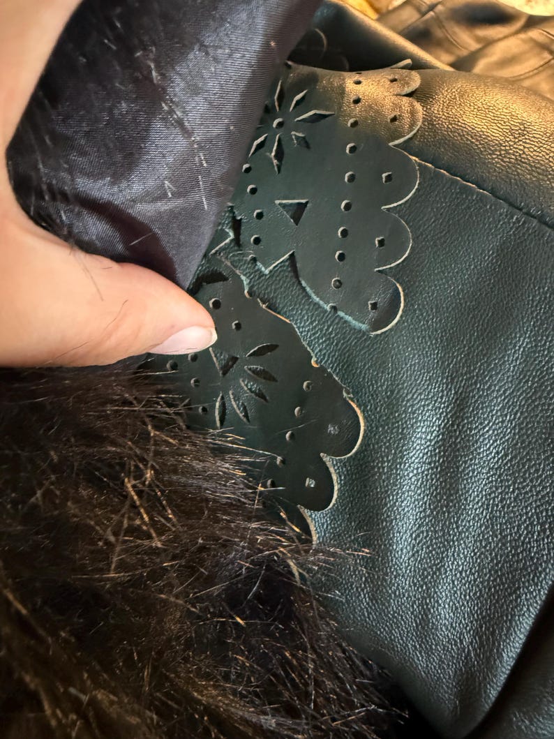 May include: Close-up of a dark green leather jacket detail. The jacket features a scalloped edge with floral cutouts. The interior lining is a dark fabric, and the collar has dark fur trim.